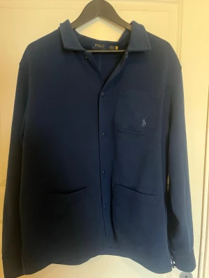 Marinblå skjorta från Polo Ralph Lauren - Stilren marinblå skjorta från Polo Ralph Lauren med klassisk krage, knappar framtill och tre fickor. Skjortan har lång ärm och diskret logga broderad på bröstfickan. Perfekt för en clean och avslappnad look.