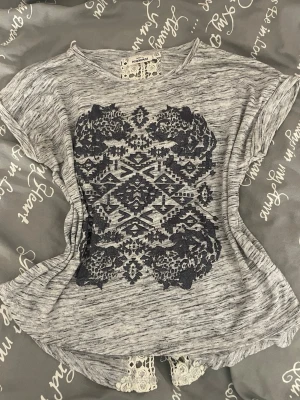 Grå topp med spetsdetaljer från KappAhl - Säljer en grå topp från KappAhl med svart broderat mönster framtill och vit spets längs ryggen. Toppen har korta ärmar och en lös passform, perfekt för en avslappnad stil. Materialet känns mjukt och toppen har en unik look med sina detaljer.