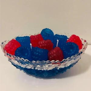 Skål med bärformade ljus - Säljer en dekorativ glasskål fylld med bärformade ljus i rött och blått. Ljusen har en tydlig hallonform och veken syns på flera av dem. Skålen är genomskinlig med ett mönstrat kant och botten. Perfekt som färgglad inredningsdetalj.