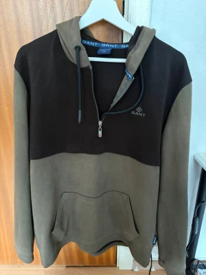 Gant Hoodie XXL - Säljer en snygg hoodie från GANT i två färger, mörkbrun upptill och olivgrön nedtill. Hoodien har dragkedja vid halsen, justerbar huva med snören och en stor magficka. GANT-logga broderad på bröstet. Perfekt för en chill och avslappnad stil. storlek XXL