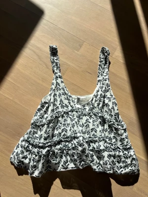 Blommig linnetopp från Pepe Jeans - Söt vit linnetopp från Pepe Jeans med svart blommigt mönster och volangdetaljer. Toppen är ärmlös med breda axelband och har en luftig, loose passform. Perfekt för varma dagar och enkel att styla med jeans eller kjol.