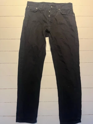 Svarta jeans från Nudie Jeans - Säljer ett par svarta jeans från Nudie Jeans med klassisk femficksmodell och raka ben. Jeansen har knappgylf och diskret logga på bakfickan. Materialet är mjukt och följsamt, perfekt för en avslappnad stil.