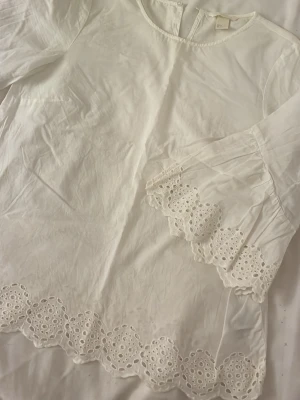 Vit blus med broderade detaljer från H&M - Säljer en vit blus från H&M med vackra broderade spetsdetaljer längs ärmslut och nederkant.