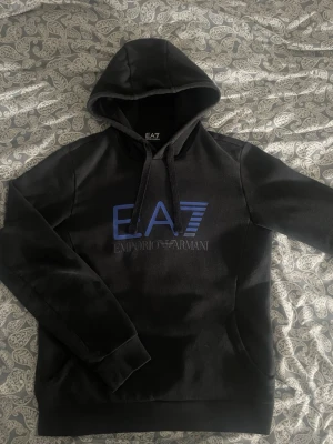 EA7 Emporio Armani hoodie - svart - strl S -  Säljer en stilren svart hoodie från EA7 Emporio Armani. Väldigt snygg och bekväm, passar till allt. Trycket framtill i blått är i fint skick utan sprickor. ✔️ Storlek: S ✔️ Skick: Mycket bra ✔️ Färg: Svart med blå logga ✔️ Inga hål eller fläckar  Perfekt hoodie för både vardag och träning