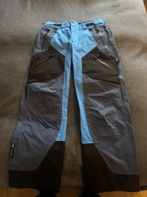 Blå och grå skidbyxor med dragkedjor - Säljer ett par täckbyxor i blått och grått med svarta detaljer. Byxorna har flera dragkedjeförsedda fickor och förstärkta partier vid knän och benslut. Materialet är slitstarkt och vattenavvisande, perfekt för vinterns äventyr. Snygg, sportig design med kontrastpaneler. Storlek S men passar M också.