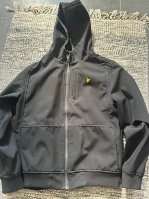 Svart jacka från Lyle & Scott - Cool svart jacka från Lyle & Scott med dragkedja och huva. Märkets ikoniska gula örn-logga på bröstet. Mjuk fleece på insidan och praktiska fickor framtill. Perfekt för dig som vill ha en stilren och bekväm jacka till vår/sommar.