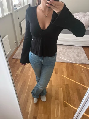 Svart blus med djup v-ringning - Snygg svart blus med djup v-ringning och långa ärmar. Modellen har utsvängda ärmslut och knytband i ryggen för en figurnära passform. Tillverkad i ett mjukt, stretchigt tyg som ger en bekväm känsla och sitter snyggt på kroppen.