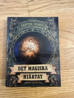 Det magiska hjärtat av Kristina Ohlsson - En snygg inbunden bok med svart och guldigt omslag, dekorerad med ett magiskt klot i centrum. Skriven av Kristina Ohlsson och utgiven av Lilla Piratförlaget. Perfekt för dig som gillar spännande och magiska berättelser.