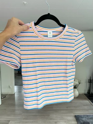 Randig croppad t-shirt, stl XS - Superfin t-shirt med färgglada ränder i blått, rosa, orange och vitt. Inköpt på barnavdelningen (skulle klassa som en XS i damstorlek). Skön och stretchig i bomullsmix, perfekt för en färgglad look. Aldrig använd!