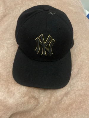 Svart New York Yankees keps - Säljer en svart keps från New York Yankees med gul broderad NY-logga framtill. Klassisk böjd skärm och justerbar baksida. Perfekt för dig som vill ha en stilren och sportig look. En fläck visas i bild 