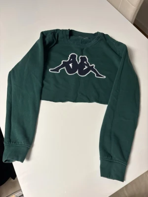 Grön croppad långärmad topp Kappa - Mörkgrön croppad långärmad topp från Kappa med stort vitt och svart logotryck framtill. Rund halsringning och mjukt bomullsmaterial. Perfekt för en sportig och trendig look.