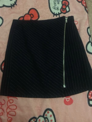 Svart kortkjol med dragkedja från H&M - Snygg svart kortkjol från H&M med tunna vita ränder och en cool snedställd dragkedja framtill. Kjolen har en stilren och modern look som passar perfekt till olika outfits. Materialet känns mjukt och bekvämt.