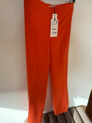 Orange byxa från Zara, XS - Snygga orange byxor från Zara i storlek XS. Byxorna har en rak passform och är tillverkade i ett slätt, vävt material. Perfekta för dig som vill sticka ut med färg och gillar trendiga, stilrena plagg. Kavajen finns till. 