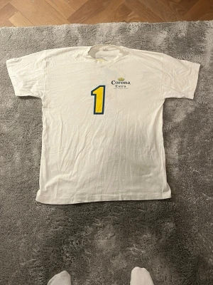 Vit Corona Extra t-shirt med tryck - Säljer en vit t-shirt från Corona Extra med stort gult och blått nummer 1-tryck på både fram- och baksidan. På ryggen finns även en tecknad figur och en fotboll med Corona-loggor. Klassisk passform och korta ärmar. Perfekt för dig som gillar unika tryck och sportig stil.