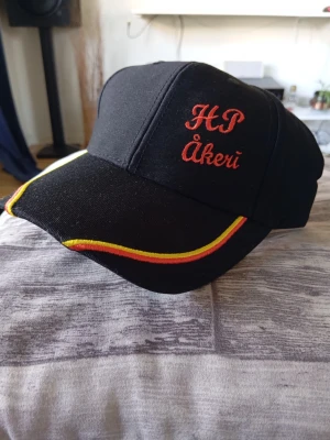 Ny Svart keps HP Åkeri brodyr - Snygg svart keps med röd brodyrtext 'HP Åkeri' framtill. Skärmen har dekorativa gula och röda linjer längs kanten. Klassisk form med böjd skärm och justerbar baksida. Perfekt för dig som gillar unik och personlig stil.