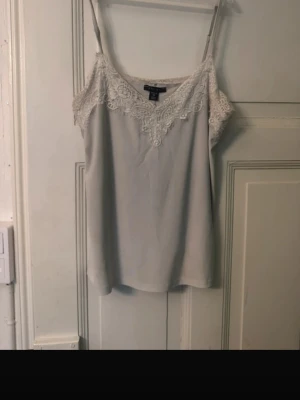 Grå spetslinnetopp från Amisu - Säljer en grå linnetopp från Amisu med tunna axelband och vacker spetsdetalj längs v-ringningen. Toppen har en mjuk och lätt känsla, perfekt för lager på lager eller sommarens varma dagar.