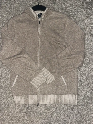 Beige stickad hoodie med dragkedja - Mysig beige stickad kashmir hoodie från boggi milano med dragkedja framtill och två fickor. Tröjan har ribbade muddar vid ärmslut och nederkant samt en justerbar huva. Perfekt för en avslappnad och stilren look.