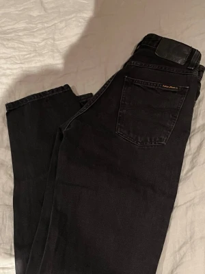 Svarta jeans från Nudie Jeans - Säljer ett par svarta jeans från Nudie Jeans med klassisk femficksmodell  Jeansen har en svart läderpatch bak i midjan och är tillverkade i kraftig denim. Perfekta för en clean och stilren look.