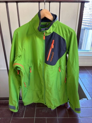 Grön vind/flecejacka från Norrøna - Snygg grön vindjacka från Norrøna med orange detaljer och svart bröstpanel. Jackan har flera dragkedjeförsedda fickor, hög krage och justerbara ärmslut. Perfekt för utomhusaktiviteter och äventyr. Tillverkad i slitstarkt syntetmaterial.