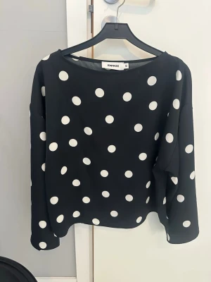 Svart prickig blus från Kappahl - Snygg svart blus från Kappahl med stora vita prickar. Blusen har rund halsringning och långa ärmar. Tillverkad i ett mjukt, lätt tyg som ger ett avslappnat men trendigt intryck. Perfekt för dig som gillar klassiska mönster med en modern twist.