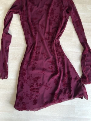 Vinröd blommig meshklänning - Vinröd kort klänning i mesh med blommigt mönster och långa ärmar. Figurnära passform och lätt transparent material ger en trendig look. Perfekt för dig som vill sticka ut med stilrena detaljer. Storlek M, men liten i storleken, relativt kort