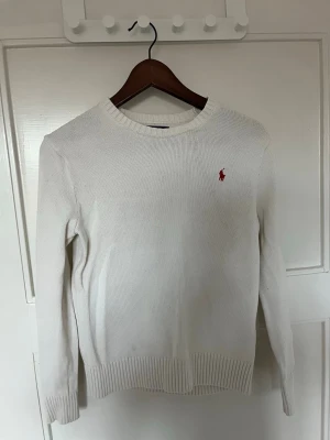 Vit stickad tröja från Ralph Lauren - Klassisk vit stickad tröja från Ralph Lauren med rund halsringning och ribbade muddar. Tröjan har den ikoniska röda Polo-loggan broderad på bröstet. Perfekt för dig som gillar stilrena och tidlösa plagg.