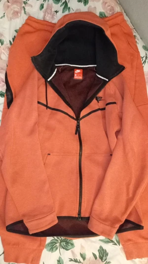 Orange/Laxrosa Nike Tech Tracksuit från Nike, M - BILLIGT! Säljer en snygg orange hoodiejacka och byxa från Nike i storlek M. Jackan har svart foder, dragkedja framtill, snörning i huvan och svarta detaljer. Perfekt för dig som gillar sportig stil och vill ha något bekvämt och coolt. Nike-logga på bröstet och på byxan