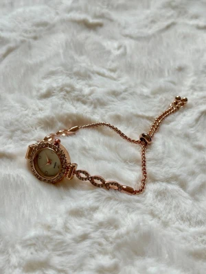 rosé Guldig klocka med strassdetaljer - Snygg rosé guldig klocka med rund urtavla och glittrande strass runt hela boetten. Länken har dekorativa detaljer och justerbar kedja med kulor i ändarna. Perfekt accessoar för dig som gillar lite extra bling.