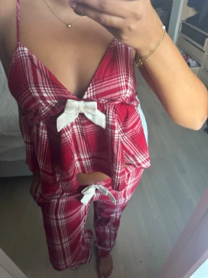 Röd- och vitrutiga pyjamas med rosetter - Säljer ett tvådelat pyjamasset i röd- och vitrutigt mönster. Linnet har smala axelband, v-ringning och en stor vit rosett framtill. Byxorna har samma rutmönster och en vit rosett i midjan. Perfekt för mysiga kvällar hemma. Från Gina tricot. Toppen är i storlek xs och byxorna i storlek 170