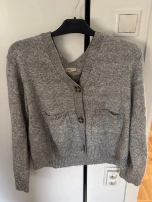 Grå kofta med fickor och knappar - Mysig grå kofta i mjukt stickat material med två stora framfickor och två knappar framtill. Koftan har v-ringning, ribbade muddar och en avslappnad passform. Perfekt att slänga över en t-shirt för en chill look.