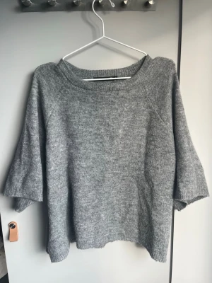 Grå stickad tröja med trekvartsärm - Mysig grå stickad tröja med bred halsringning och trekvartsärmar. Tröjan har en avslappnad passform och ribbade kanter vid ärmslut och nederkant. Perfekt för lager på lager och enkel att matcha med olika outfits. Hör av er innan köp, brukar svara inom nån timme.