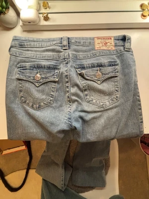 Ljusblå True Religion jeans - Säljer ett par ljusblå jeans från True Religion med ikoniska ficklock och dekorativa sömmar bak. Jeansen är bootcut, klassisk femficksmodell och silverfärgade knappar. Materialet är mjukt denim i bomull. Perfekta för en avslappnad och trendig look. Dem är även uppsydda i benen men går att sprätta upp.