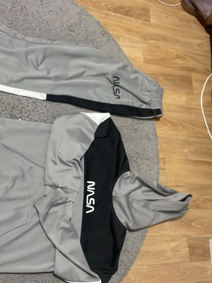 Grå NASA loungewear set - Snyggt loungewear set från NASA i grått och svart med vita detaljer. Setet består av en hoodie med ficka framtill och matchande byxor med resår i midjan. NASA-loggan syns tydligt på både tröjan och byxan. Perfekt för chill dagar hemma eller när du vill ha en avslappnad stil.
