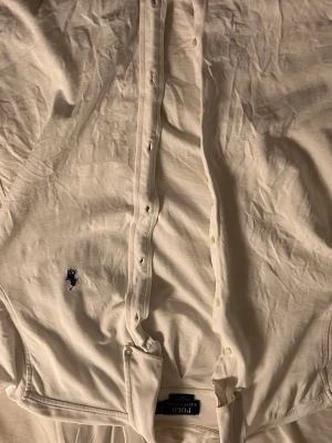 Vit skjorta från Polo Ralph Lauren - Klassisk vit skjorta från Polo Ralph Lauren med blå broderad logga på bröstet. Skjortan har knappar framtill, lång ärm och normal passform. Tillverkad i bomull, perfekt till både jeans och chinos för en clean look. Modellen är 181cm lång och väger 80 kg. Sitter ganska tajt på mig. Men passar perfekt för någon runt 177 cm lång.