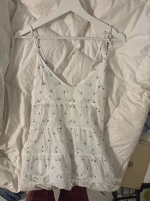Vit linnetopp med blått mönster - Söt vit linnetopp med tunna axelband och små blå broderade detaljer. Toppen har volang nedtill och smockad rygg för extra komfort. Perfekt för varma dagar och enkel att matcha med jeans eller kjol.