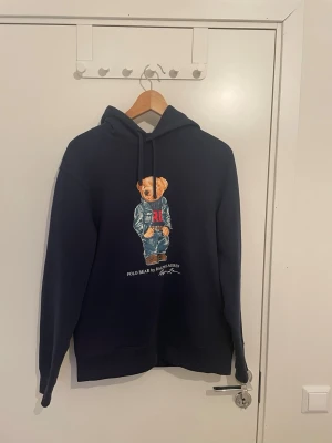 Mörkblå hoodie Polo Ralph Lauren - Mörkblå hoodie från Polo Ralph Lauren med ikoniska Polo Bear-trycket på bröstet. Klassisk huva med dragsko och långärmad modell. Tillverkad i mjuk bomull för maximal komfort. Perfekt för en avslappnad och stilren look. Självklart äkta. Kan gå ner i pris vid snabb affär