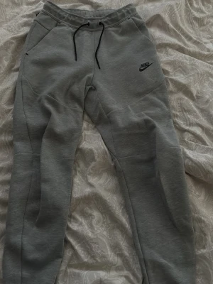 Gråa mjukisbyxor från Nike - Snygga gråa mjukisbyxor från Nike med svart logga på vänster ben. Byxorna har dragsko i midjan, två sidofickor fram och en ficka med dragkedja bak. Tillverkade i mjukt och bekvämt material med smal passform och ribbade benslut.