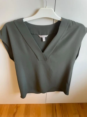Mörkgrön blus med v-ringning - Stilren mörkgrön blus med bred v-ringning och avslappnad passform. Blusen har korta ärmar och är tillverkad i ett mjukt, lätt material som ger ett snyggt fall. Perfekt att matcha med både jeans och kjol.