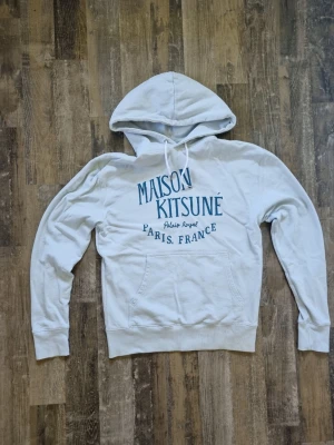 Maison Kitsuné blå hoodie - Snygg blå hoodie från Maison Kitsuné med blå texttryck på bröstet där det står 'Maison Kitsuné Paris, France'. Klassisk känguruficka framtill, ribbade muddar och dragsko i huvan. Tillverkad i mjuk bomull för en skön och avslappnad look.