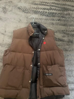 Brun dunväst från Canada Goose - Canada goose väst, bra skicl förutom flärpen på dragkedjan som är borta, storlek XXL men passar mig som är M helt ok så ska passa på L