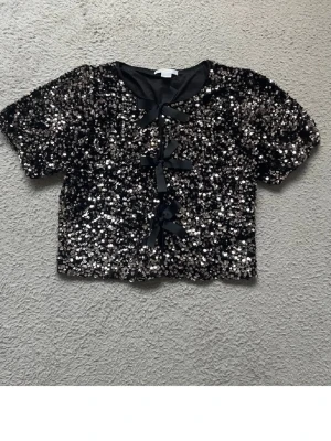 Svart paljettopp med rosetter - Helt oanvänd topp från Gina tricot, nypris ligger på 350kr, använd gärna köp nu!!🩷🩷Glittrig svart topp täckt med paljetter och dekorerad med tre stora rosetter framtill. Kortärmad modell med rund halsringning och croppad passform. Perfekt för dig som vill sticka ut och glänsa.