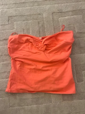 Korallfärgad bandeau topp - Säljer en snygg korallfärgad bandeau topp med twistad detalj framtill. Toppen är axelbandslös och har en tight passform som framhäver figuren. Perfekt till sommaren och enkel att matcha med shorts eller kjol.