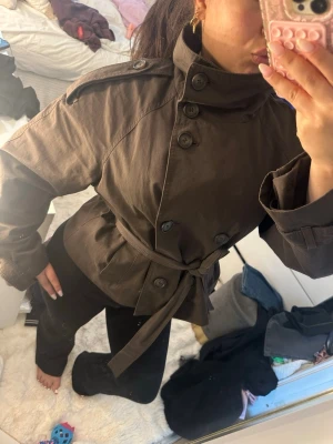 Brun trenchcoat med bälte - Snygg brun trenchcoat med hög krage, dubbelknäppning och knytskärp i midjan. Jackan har axelklaffar och stora knappar framtill. Perfekt för dig som vill ha en trendig och stilren look. Materialet känns slitstarkt och jackan har en kortare, boxig passform. Jackan har en liten missfärgning från av att jag strykt den