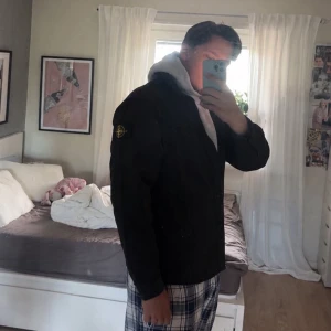 Svart overshirt från Stone Island - Säljer en svart overshirt från Stone Island med klassisk krage och knappstängning framtill. Jackan har två bröstfickor med knappar och den ikoniska Stone Island-loggan på vänster ärm. Perfekt för lager-på-lager och streetwear.