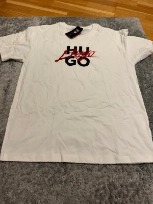 Vit HUGO t-shirt med tryck - Snygg vit t-shirt från HUGO med svart och rött tryck framtill där loggan är överstruken med röd text. Klassisk passform och rund halsringning. Perfekt till jeans eller shorts för en clean och modern look.