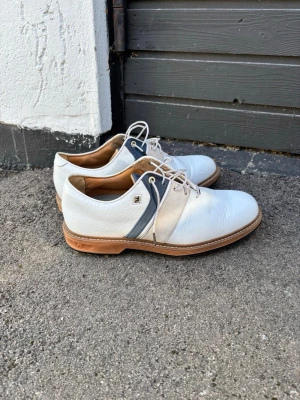 Vita FootJoy sneakers med blå detalj - Säljer ett par vita golfskor från FootJoy med blå och grå detaljer på sidan. Skorna har snörning, rund tå och en klassisk platt sula. Tillverkade i skinn. storlek 41 men passar 42, 27 cm innermått