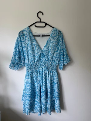 Blåvit kortklänning från New Yorker - Söt blåvit kortklänning från New Yorker med v-ringning, tajt i midjan och vida korta ärmar. Klänningen har ett blommigt mönster och är tillverkad i ett lätt material som passar perfekt till varma dagar.