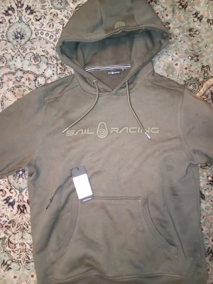Olivgrön hoodie från Sail Racing - Olivgrön hoodie från Sail Racing med stor logga på bröstet och klassisk huva med dragsko. Tröjan har en stor magficka och ribbade muddar. Materialet är mjukt och bekvämt, perfekt för en avslappnad stil.förlåt för dålig kvalitè då jag tappade min mobil annars är den fräsch och osnvänd med tags