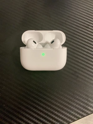 Apple AirPods Pro trådlösa hörlurar - Säljer ett par vita Apple AirPods Pro med laddningsetui. Hörlurarna är in-ear och har en stilren, kompakt design i blank plast. Perfekta för dig som vill ha trådlös frihet och bra ljudkvalitet. Laddningsetuiet har en LED-indikator på framsidan.