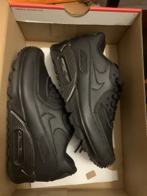 Nike Air Max 90 helsvarta sneakers - Snygga Nike Air Max 90 sneakers i helsvart färg med klassisk siluett. Skorna har ovandel i läder och textil, synlig Air Max-dämpning i sulan och diskreta Nike-loggor på sidorna. Perfekta för dig som gillar en clean och stilren look.